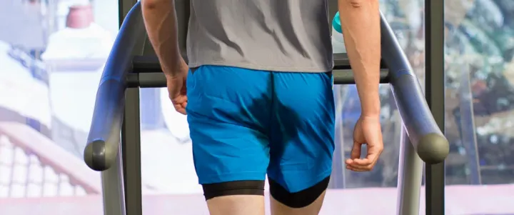 A runner on a treadmill with a sweat-darkened patch on the seat of his blue shorts.